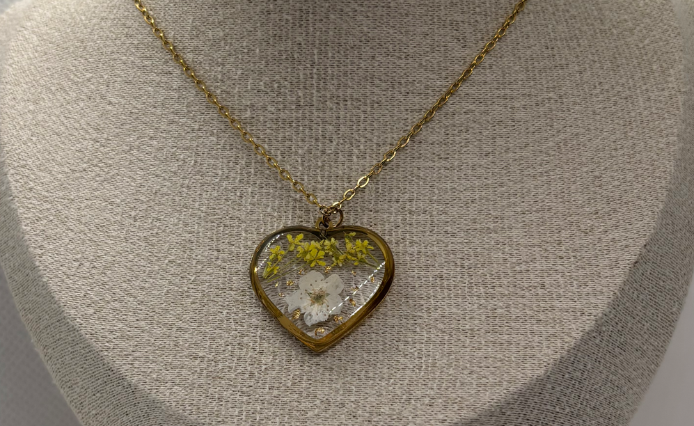 A gold heart-shaped pendant necklace with dried yellow and white flowers, displayed on a textured beige fabric jewelry bust.
